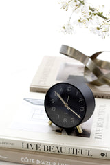 Karlsson Lofty alarm clock in matte black with gold legs and gold hands on a Live Beautiful book stack beside a silver sculptural object and white blossom branches on a white surface.