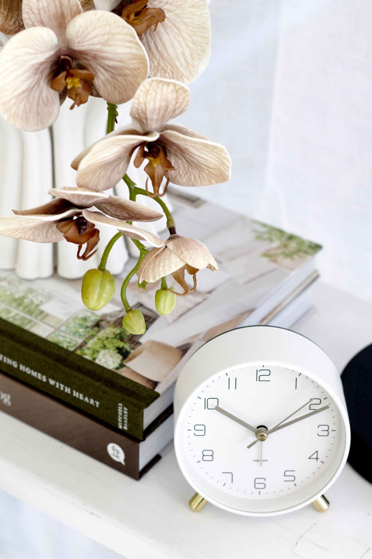 White Karlsson Lofty alarm clock on stacked books beside orchids and a lamp