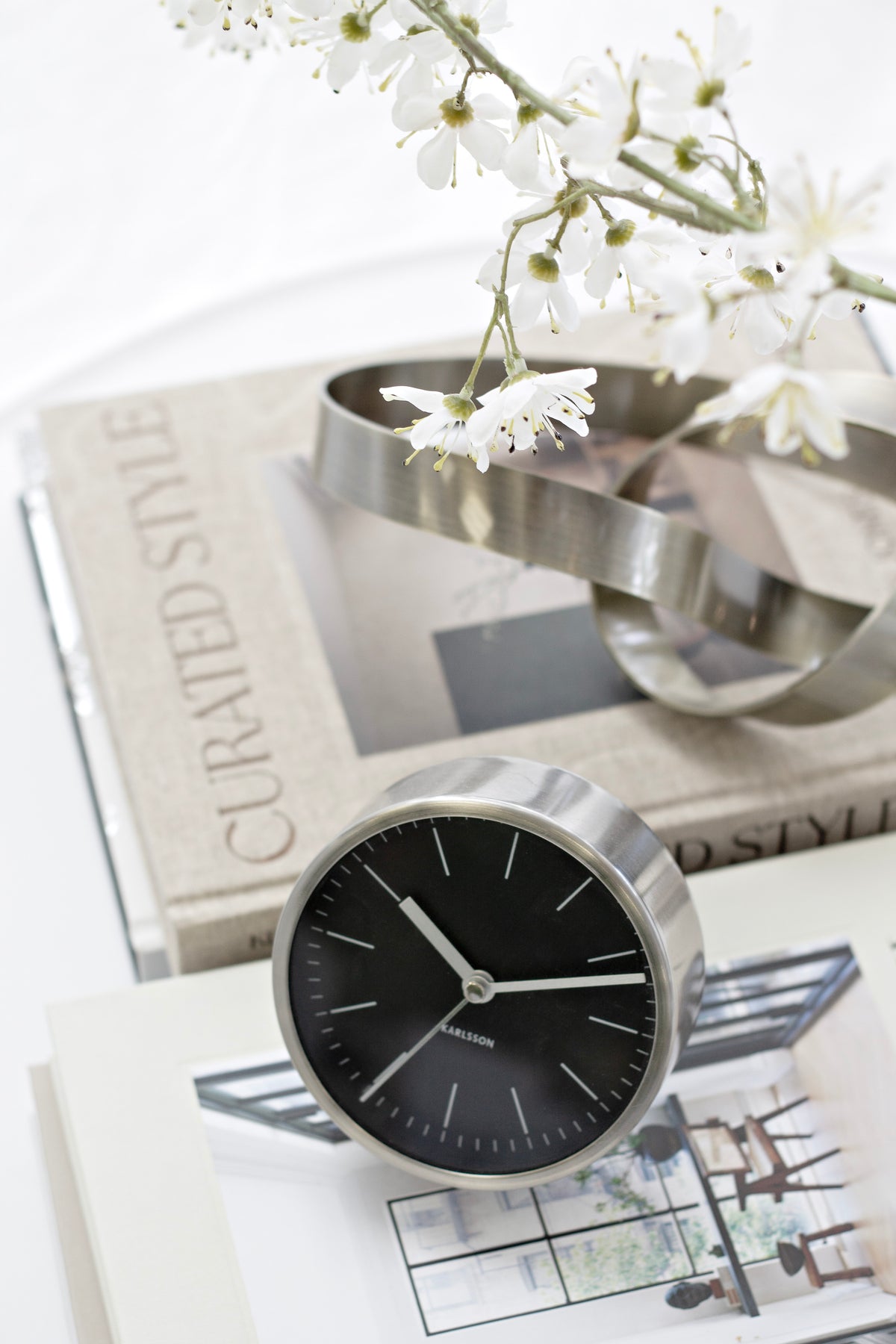 Minimal Alarm Clock with black dial on stacked books beside a chrome sculpture and white blossom