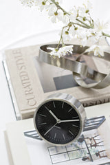 Karlsson Minimal Alarm Clock with black dial and nickel case styled on stacked Curated Style books with chrome sculptural object and white blossom branches in a white interior.