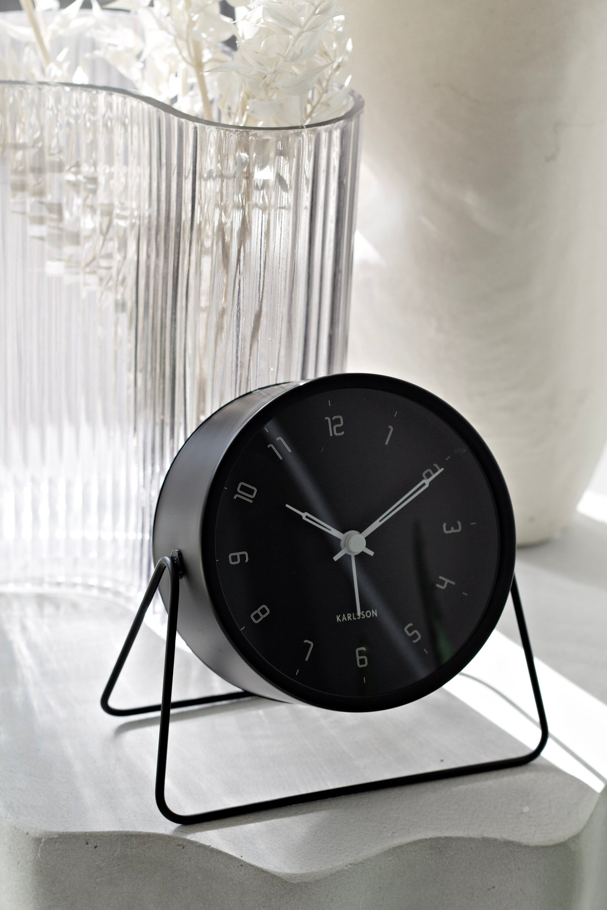 Black Stark Alarm Clock on its wire stand beside a ribbed glass vase of dried stems