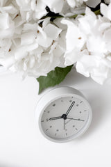 A white Karlsson Tinge alarm clock styled in a flat lay beside a lush arrangement of white hydrangeas on a clean white surface, evoking a calm, minimal bedside aesthetic.