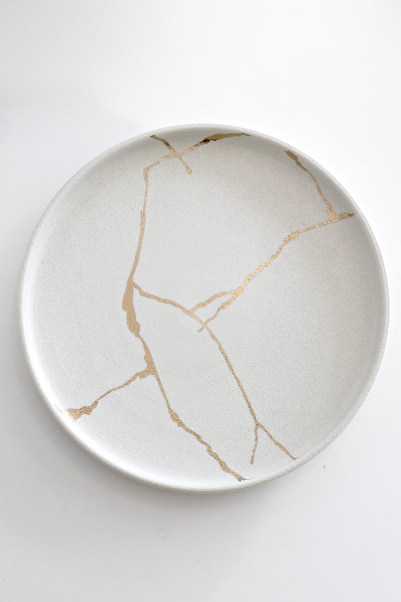 Cream porcelain plate with gold Kintsugi-inspired crack design, styled minimal and modern on white surface.