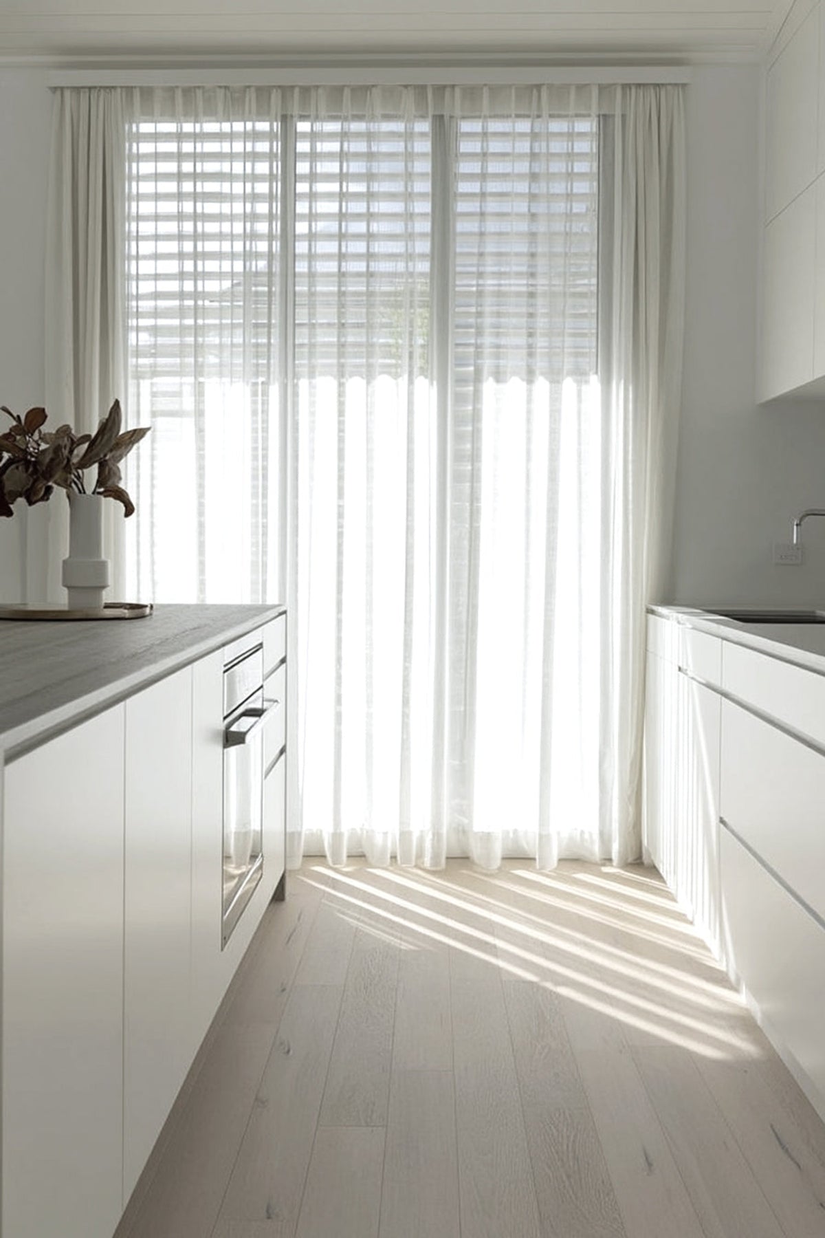 White kitchen in strong sunlight with a pale vase of leafy stems on the benchtop by a sheer curtain