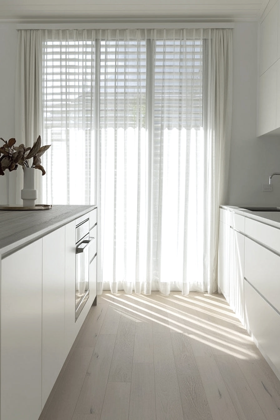 White kitchen in strong sunlight with a pale vase of leafy stems on the benchtop by a sheer curtain