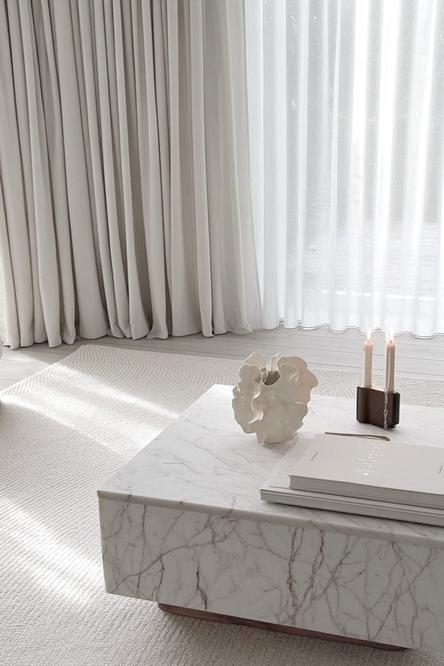 White coral vase on a marble coffee table beside a candle and stacked books in a pale living room