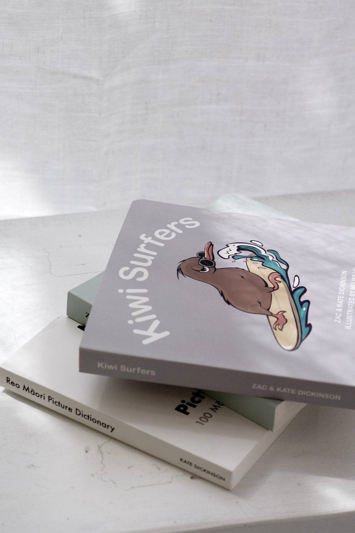 Kiwi Surfers children’s picture book with a cartoon kiwi on a surfboard, styled on a stack of books on a light stone surface