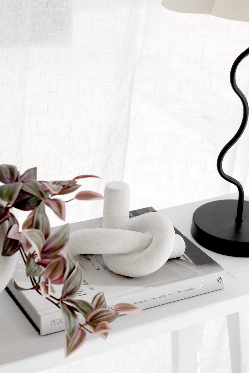 White knot ornament styled on books with plant and lamp; modern sculptural decor accessory for shelves or coffee tables.