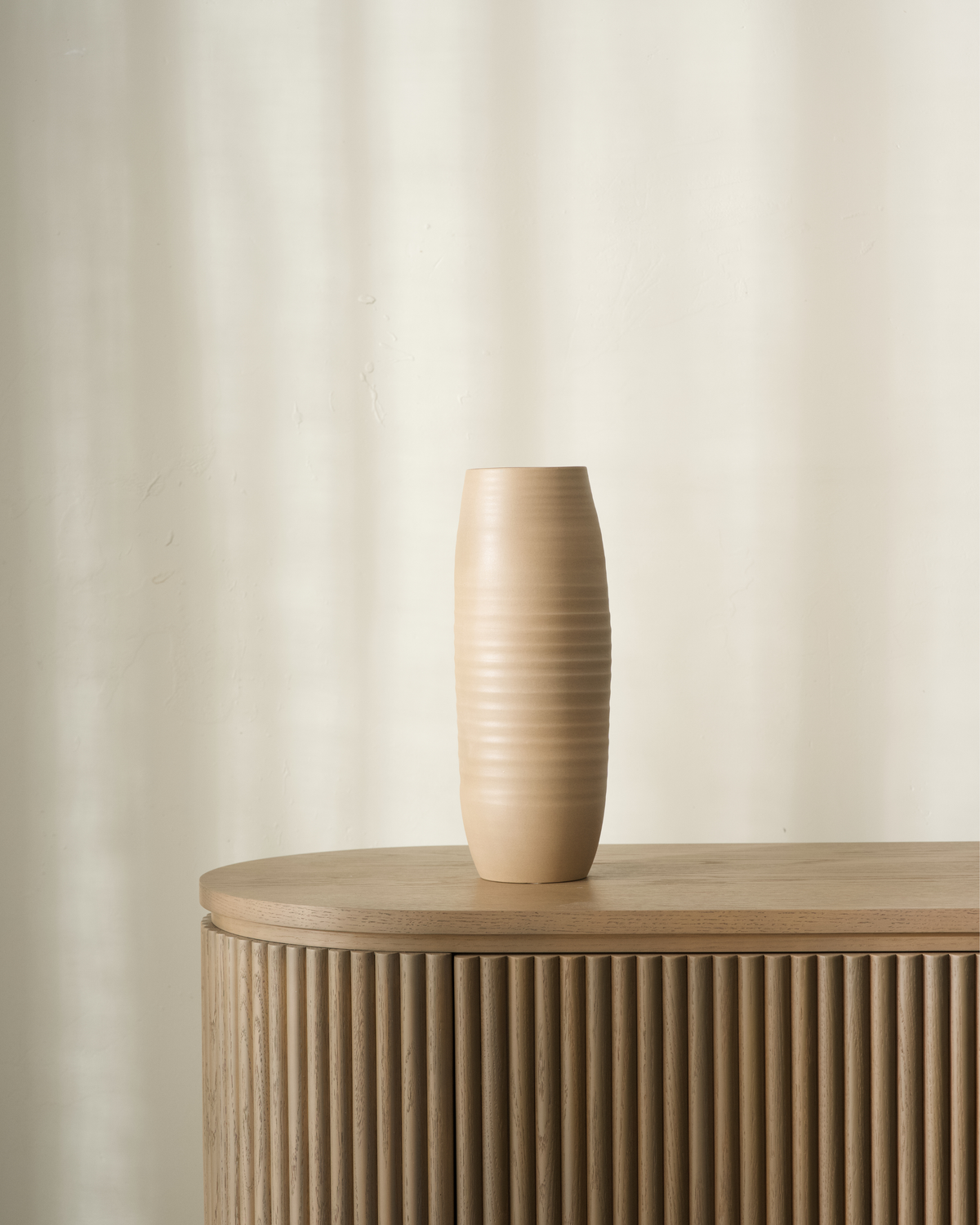 Kona Vase by Ned Collections in Small Mud, a tall ribbed sand coloured ceramic vase on a fluted oak sideboard