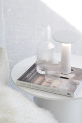 A white LED Tilt rechargeable touch lamp styled on a round white side table alongside a ribbed glass carafe, matching tumbler, and a High on Living interior design book, creating a minimal, light-filled Japandi-inspired bedside vignette.