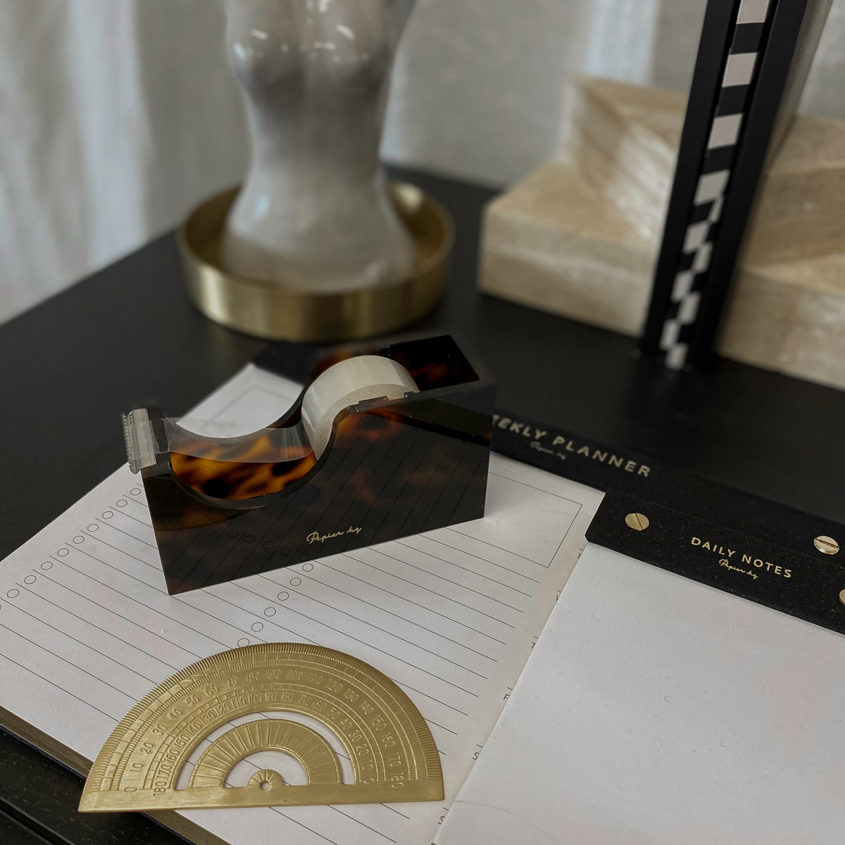 Tortoiseshell tape dispenser on a black desk with a gold notepad and vase