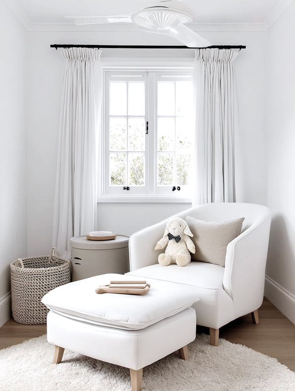 Modern white nursery corner with a cosy armchair and footstool, a plush lamb toy with a bow tie on the seat, soft neutral textiles, a knitted storage basket, and a white ceiling fan above.