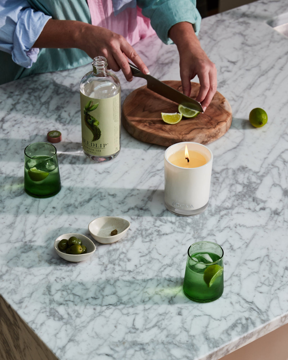 A lit Ecoya Madison candle on a marble bench while someone slices limes, with green glasses and a bottle alongside