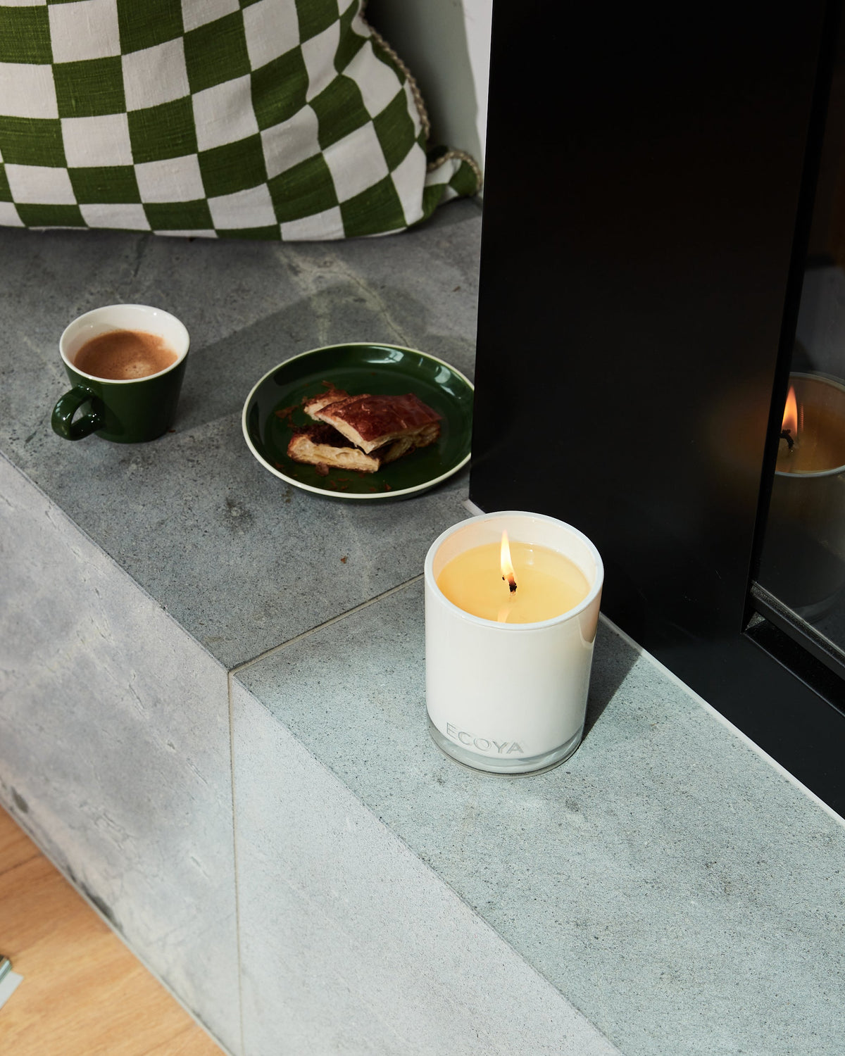 A lit Ecoya Madison candle on a concrete hearth beside a fireplace, with a green mug and plate and a checked cushion