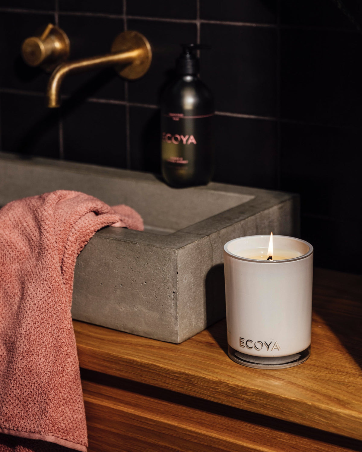 Lit Ecoya Madison candle on a timber vanity beside a concrete basin, hand wash and pink towel