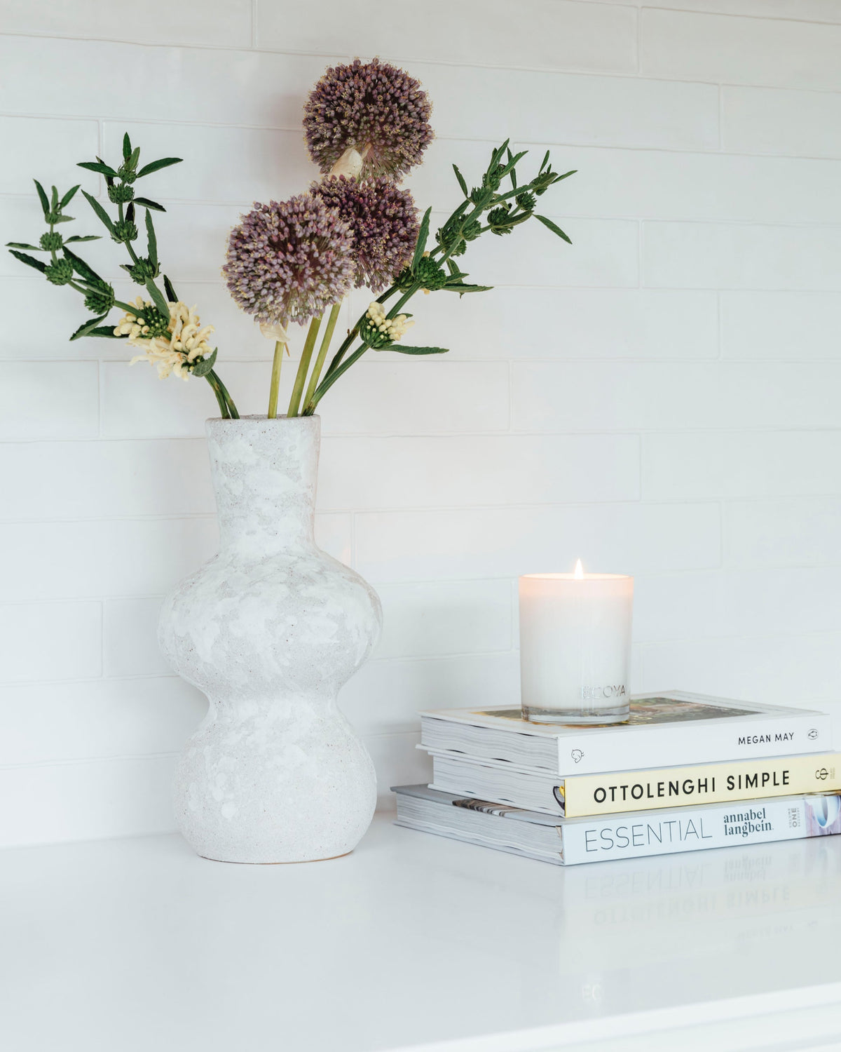 A lit Ecoya Madison candle on a stack of cookbooks beside a textured white vase of alliums