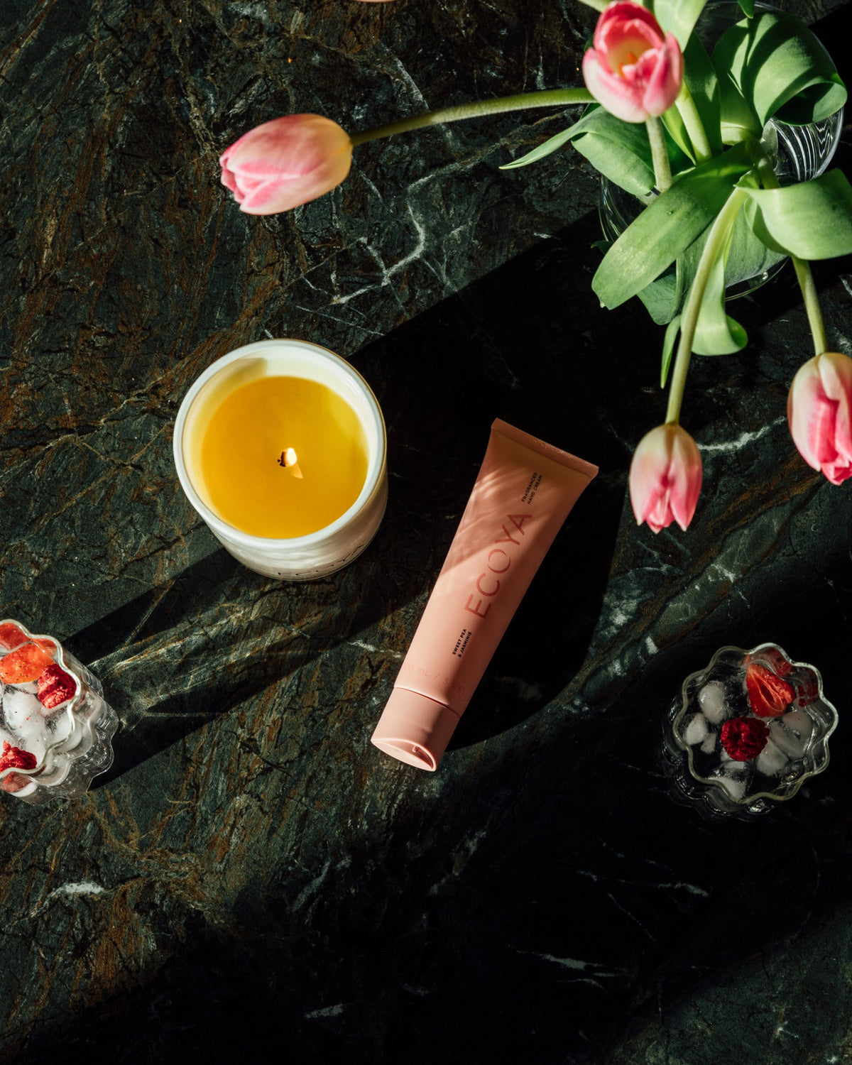 Peach Ecoya hand cream tube on green marble beside a lit candle and tulips