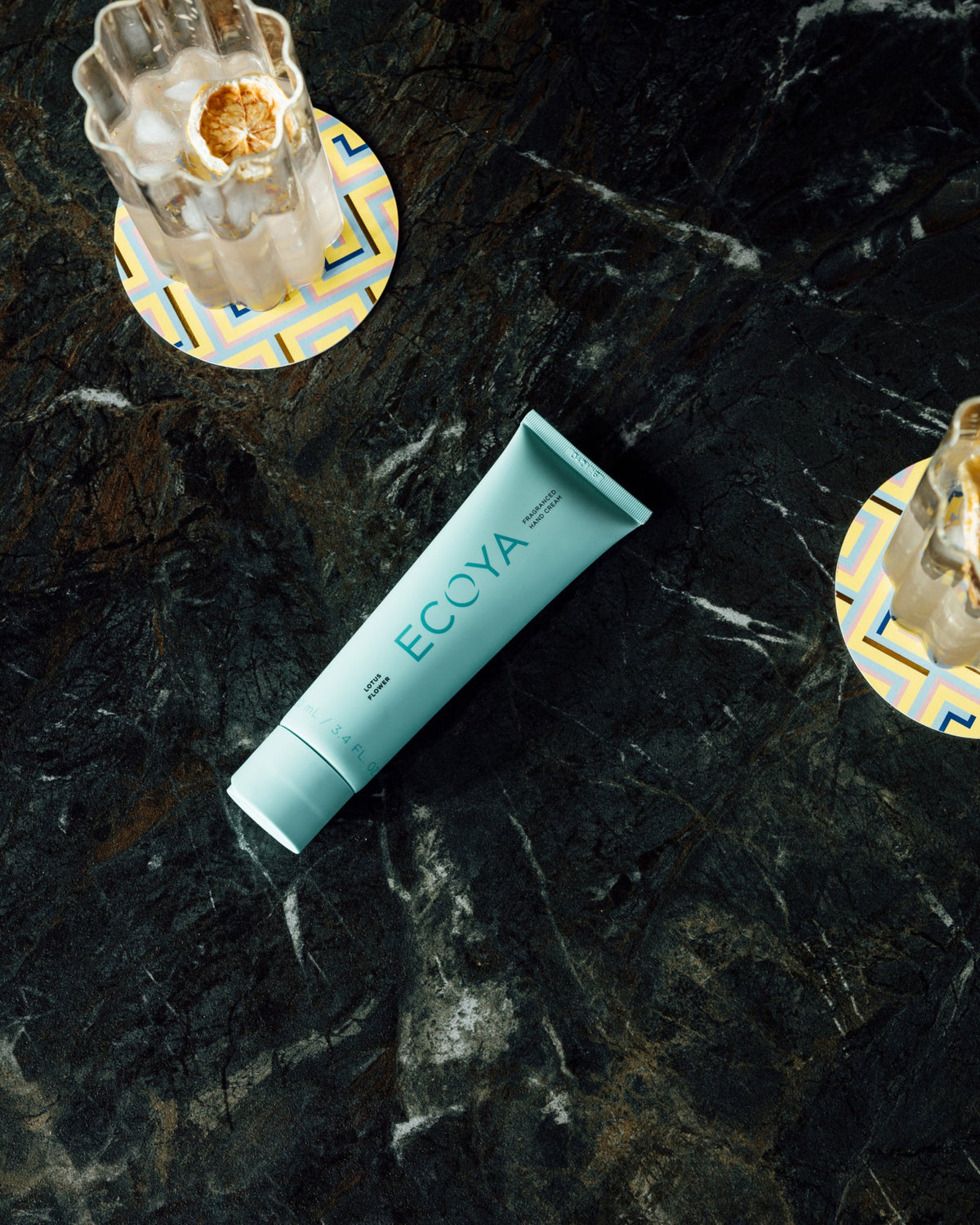 Teal Ecoya hand cream tube on dark marble beside glassware