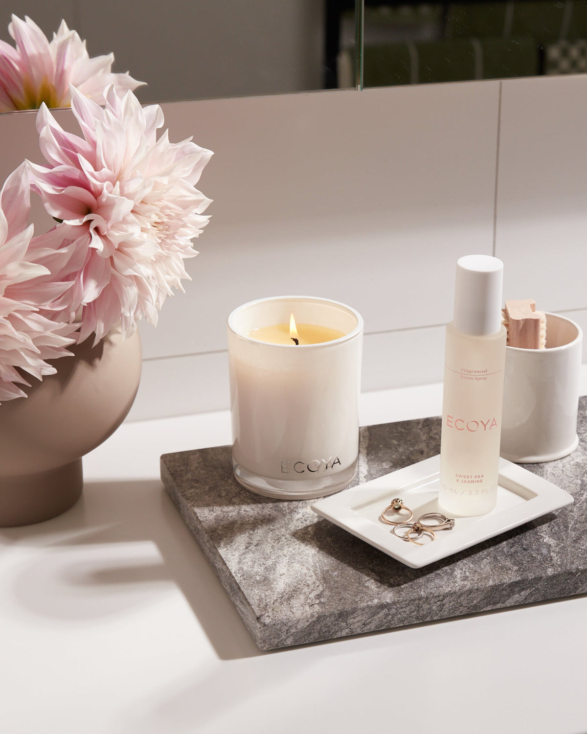 Lit Ecoya Madison candle on a stone tray with pink peonies and bathroom bottles