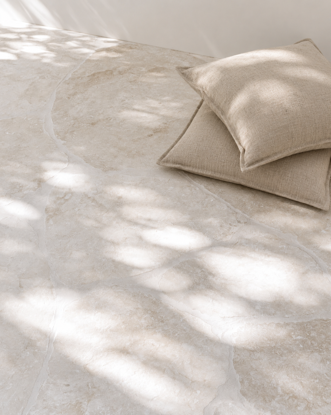 Two Linen Latte cushions with flanged edges stacked on a marble floor in dappled sunlight