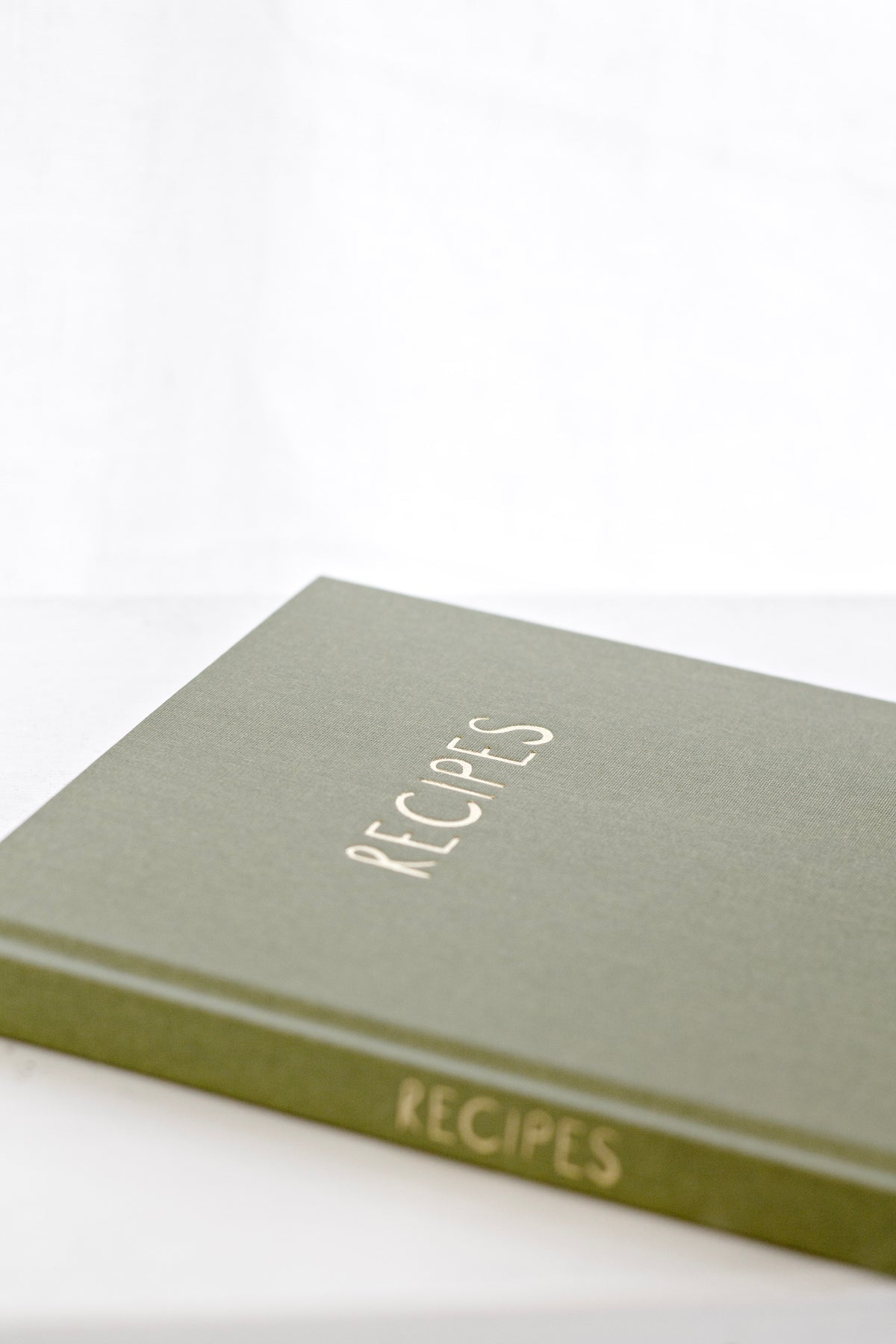 Olive linen recipe journal lying flat, angled to show the embossed title and spine