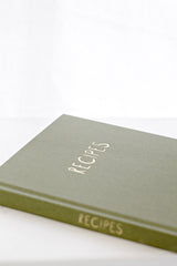 Olive green linen recipe journal with the word “RECIPES” embossed in gold on the cover, styled on a light surface in soft natural light.