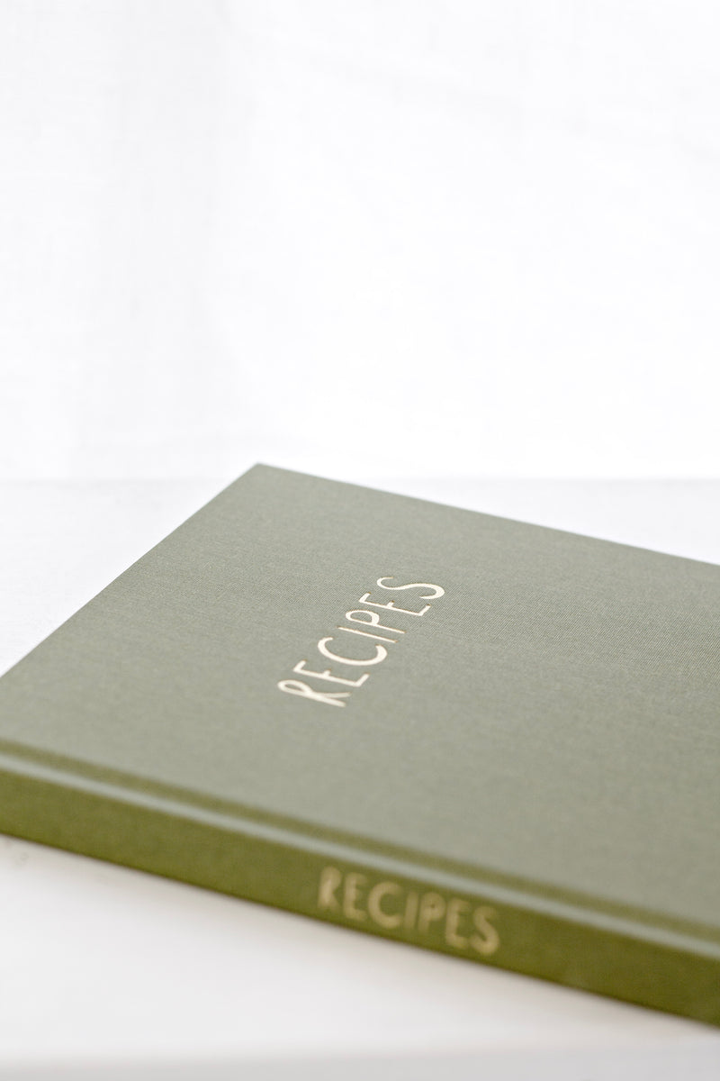 Olive green linen recipe journal with the word “RECIPES” embossed in gold on the cover, styled on a light surface in soft natural light.