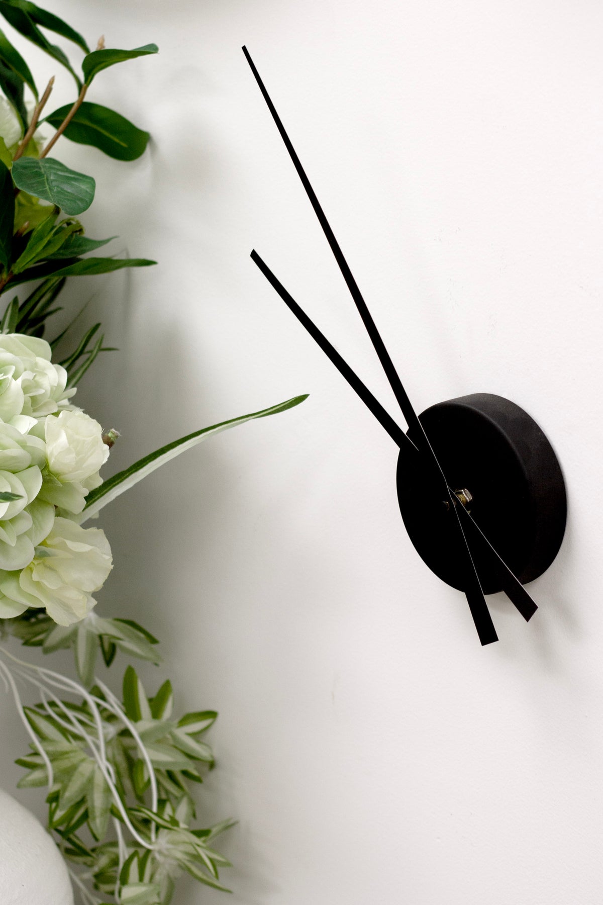 Black minimalist wall clock with floating hands mounted on white wall beside green foliage