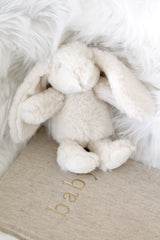 Cream plush bunny toy with floppy ears resting on a linen baby journal with gold lettering, styled on soft white faux fur