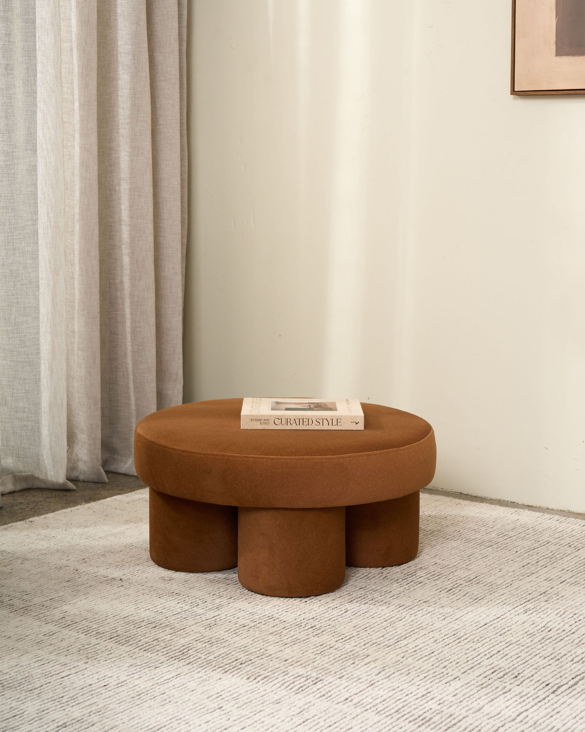 Caramel round ottoman holding the book Curated Style on a pale rug beside linen curtains