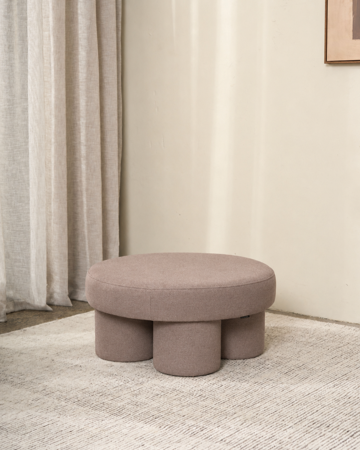 Round ottoman in a light taupe textured weave with four chunky legs, on a pale rug