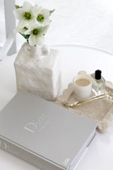 White sculptural ceramic vase with dual handles styled with flowers on a coffee table beside a Dior book and travertine tray.