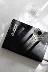 Black 2026 Made of Tomorrow A5 daily diary in onyx faux leather on a white wood surface, styled with a clear glass cup and square glass perfume bottle casting shadows.