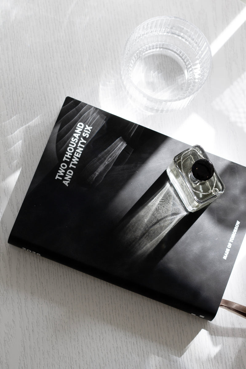 Black 2026 Made of Tomorrow A5 daily diary in onyx faux leather on a white wood surface, styled with a clear glass cup and square glass perfume bottle casting shadows.