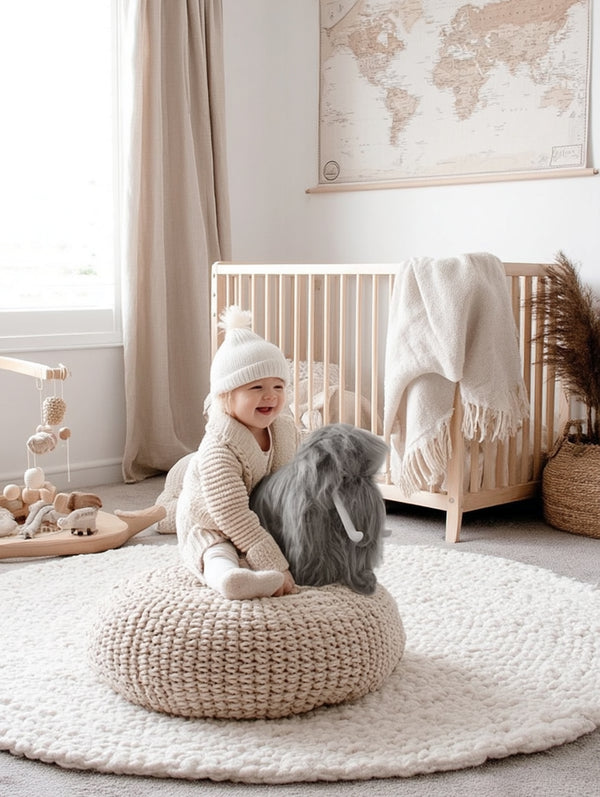 Toddler sitting on a knitted pouf in a neutral-toned nursery, smiling while holding a large plush grey mammoth toy with white tusks and long shaggy fur.