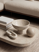 Minimalist coffee table styling featuring a solid white marble loop sculpture, marble pedestal bowl, stone sphere, and stacked neutral books beside a cream boucle sofa.