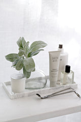 White marble-look tray styled with greenery in a glass vase, candle, hand cream, body products and wick trimmer on a white surface in soft light.