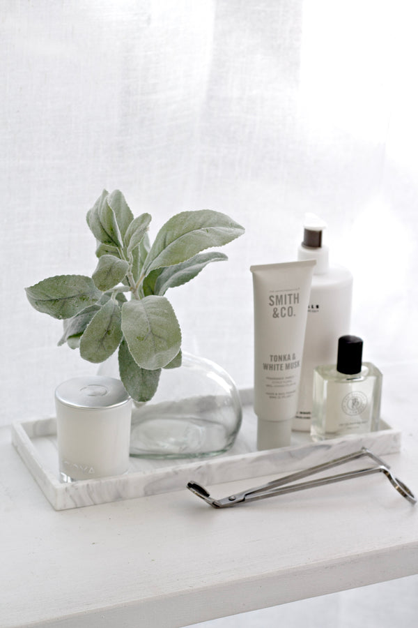 White marble-look tray styled with greenery in a glass vase, candle, hand cream, body products and wick trimmer on a white surface in soft light.