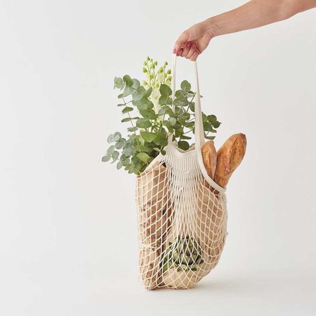 Hand lifting the cream string bag loaded with eucalyptus and a baguette against a pale wall
