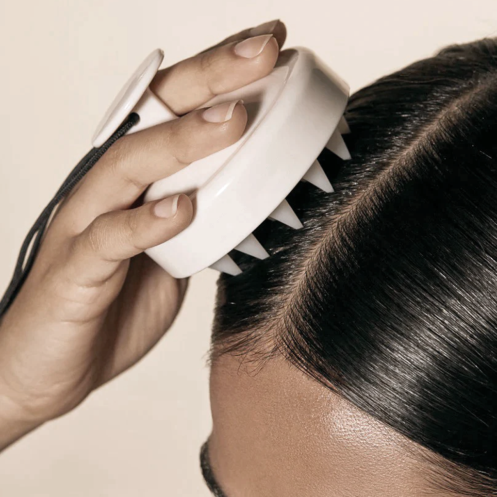 A white scalp massager being worked through dark hair at the roots, shown on a beige background