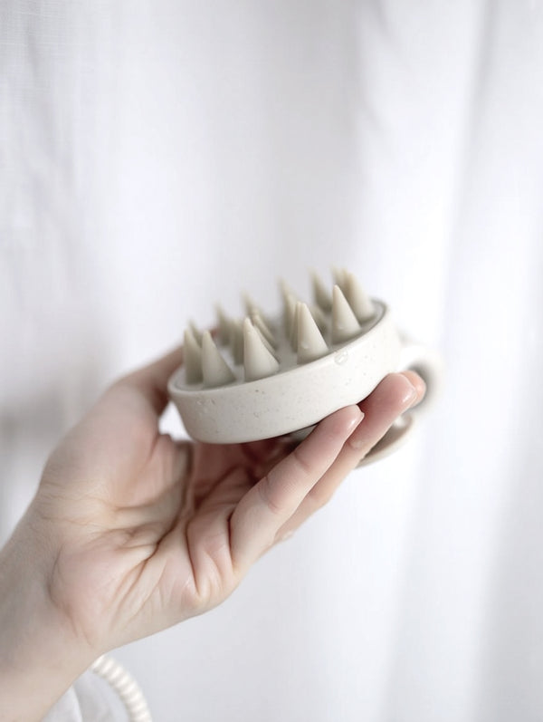 A hand holds a neutral-toned silicone scalp massager with soft bristles and a wheat straw handle, set against a soft white background.
