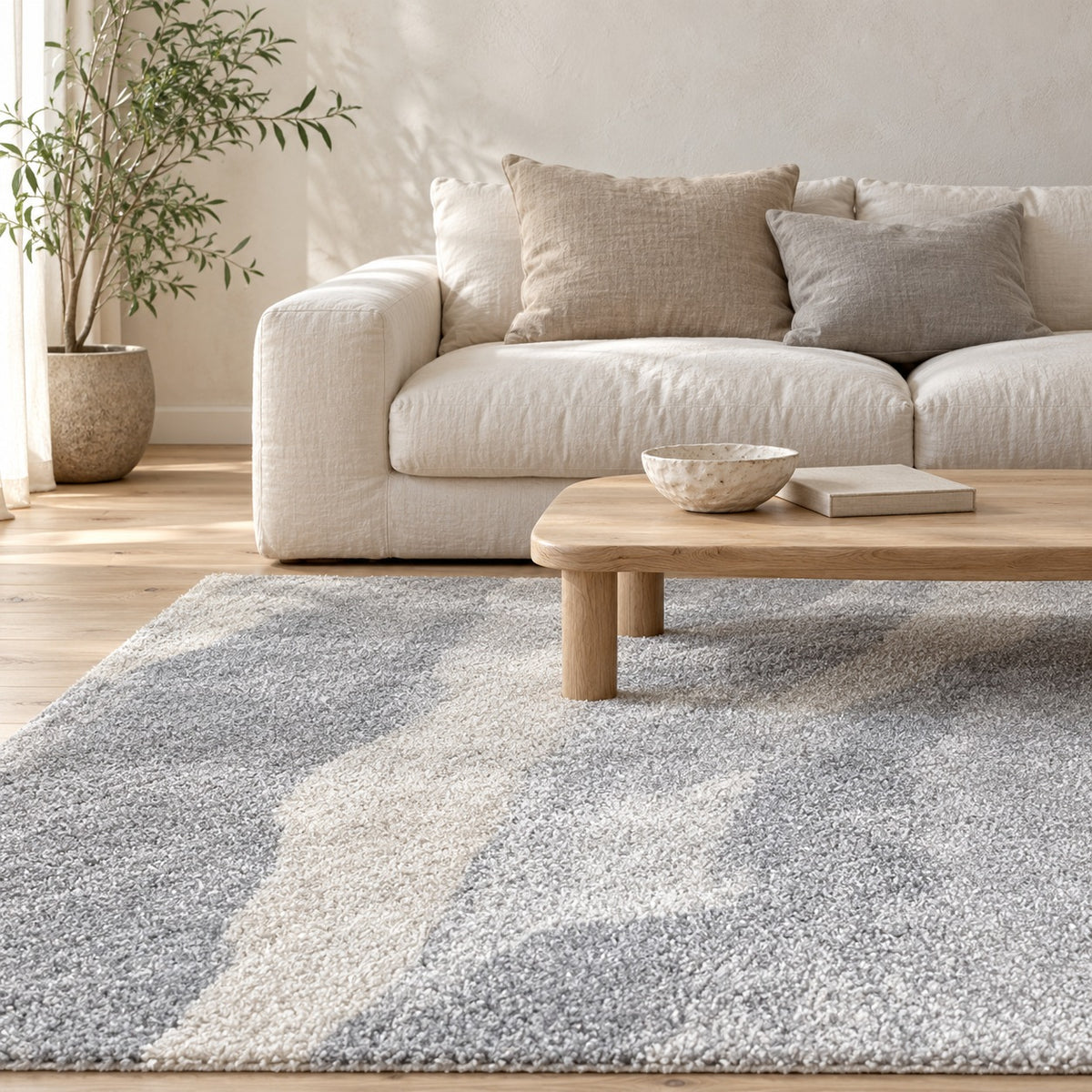 Mercer Cloud Grey Rug under a timber coffee table in front of a linen sofa, with an olive tree by the window