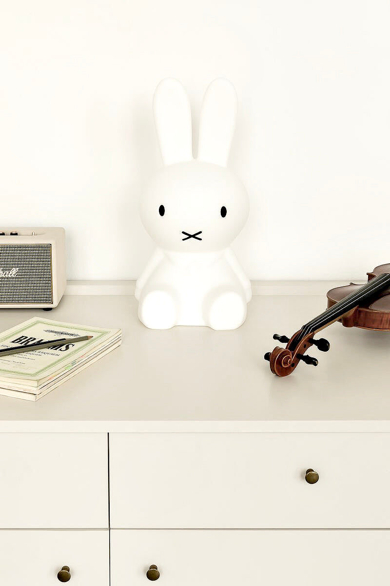 Large white Miffy bunny lamp on a light dresser between a small speaker and a violin in a neutral kids room
