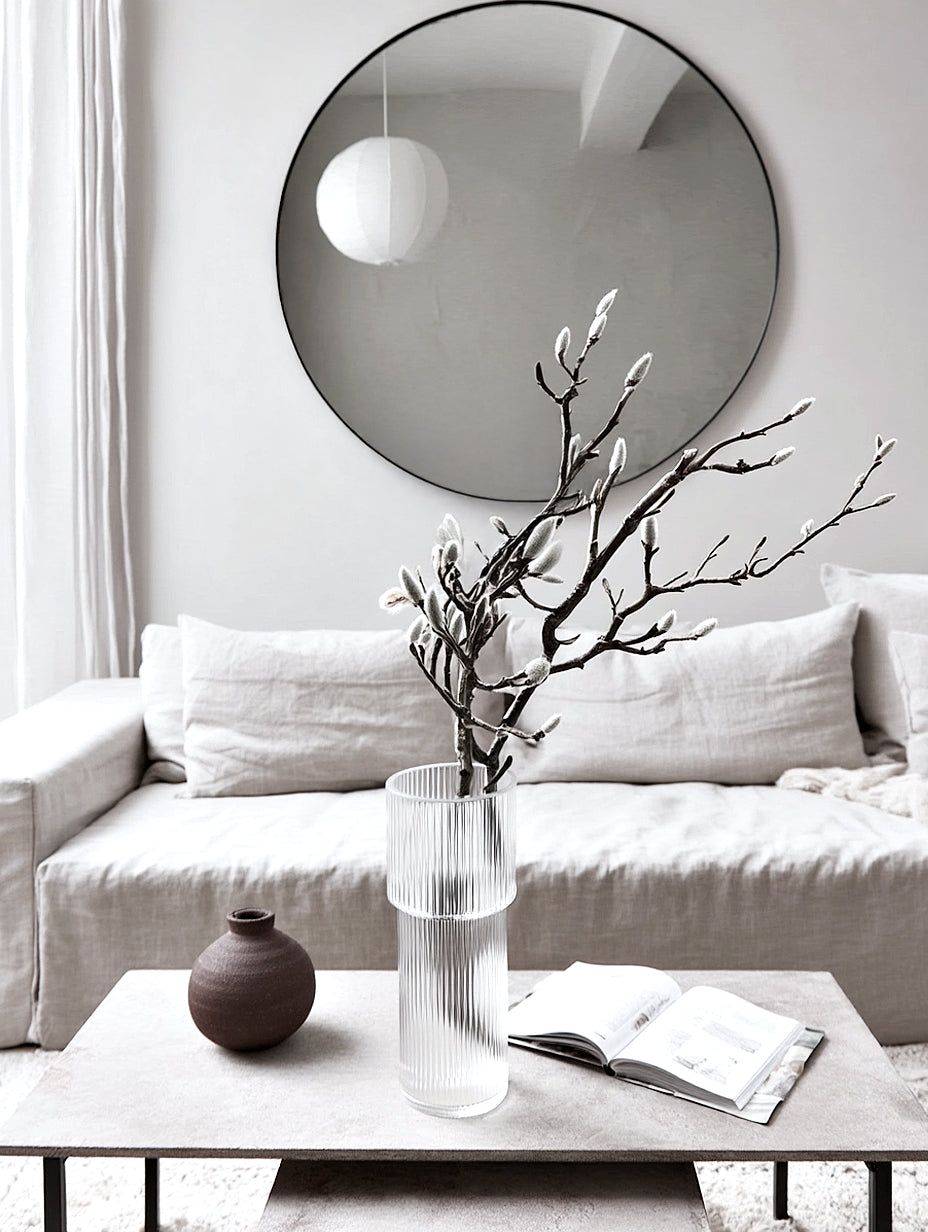 Round black frame mirror above a linen sofa, with a glass vase of branches on the coffee table