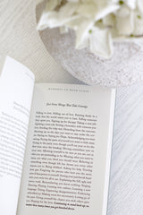 A close-up, flat-lay style image of the poetry book 'Moments to Hold Close' by Molly Burford, open to the page titled "Just Some Things That Take Courage." The book rests on a white wood surface with soft, out-of-focus white flowers in the background, creating a gentle, serene, and inspiring mood.
