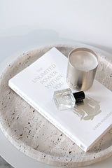 Round travertine Moon tray styled with a white book, a metallic candle and a small perfume bottle on a pale surface in a minimal neutral setting