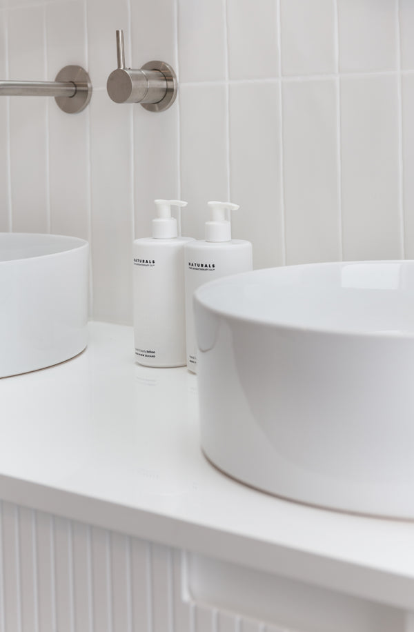 Santal & Vanilla Orchid vegan hand & body lotion styled on a white bathroom vanity with vessel basins.