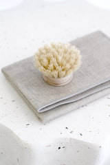 Neutral natural cotton dishcloth folded neatly with a wooden scrub brush on a speckled white benchtop in soft, minimal lighting.