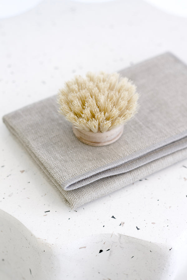 Neutral natural cotton dishcloth folded neatly with a wooden scrub brush on a speckled white benchtop in soft, minimal lighting.