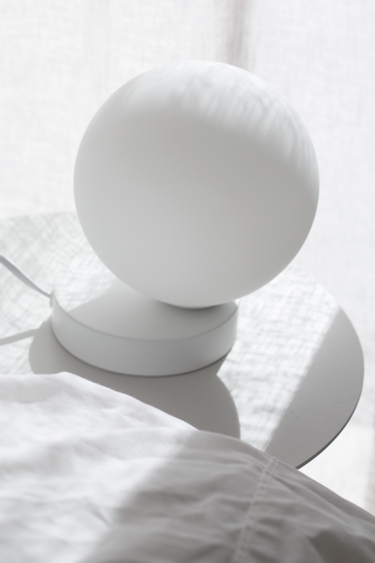 White globe table lamp on round bedside table with linen bedding in soft morning light.